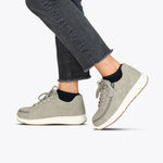 A person wearing Women's Grey Suede BILLY Comfort Plush shoes by BILLY Footwear, with white soles and black socks, pairs them with frayed-hem dark jeans while stepping forward against a plain light background.