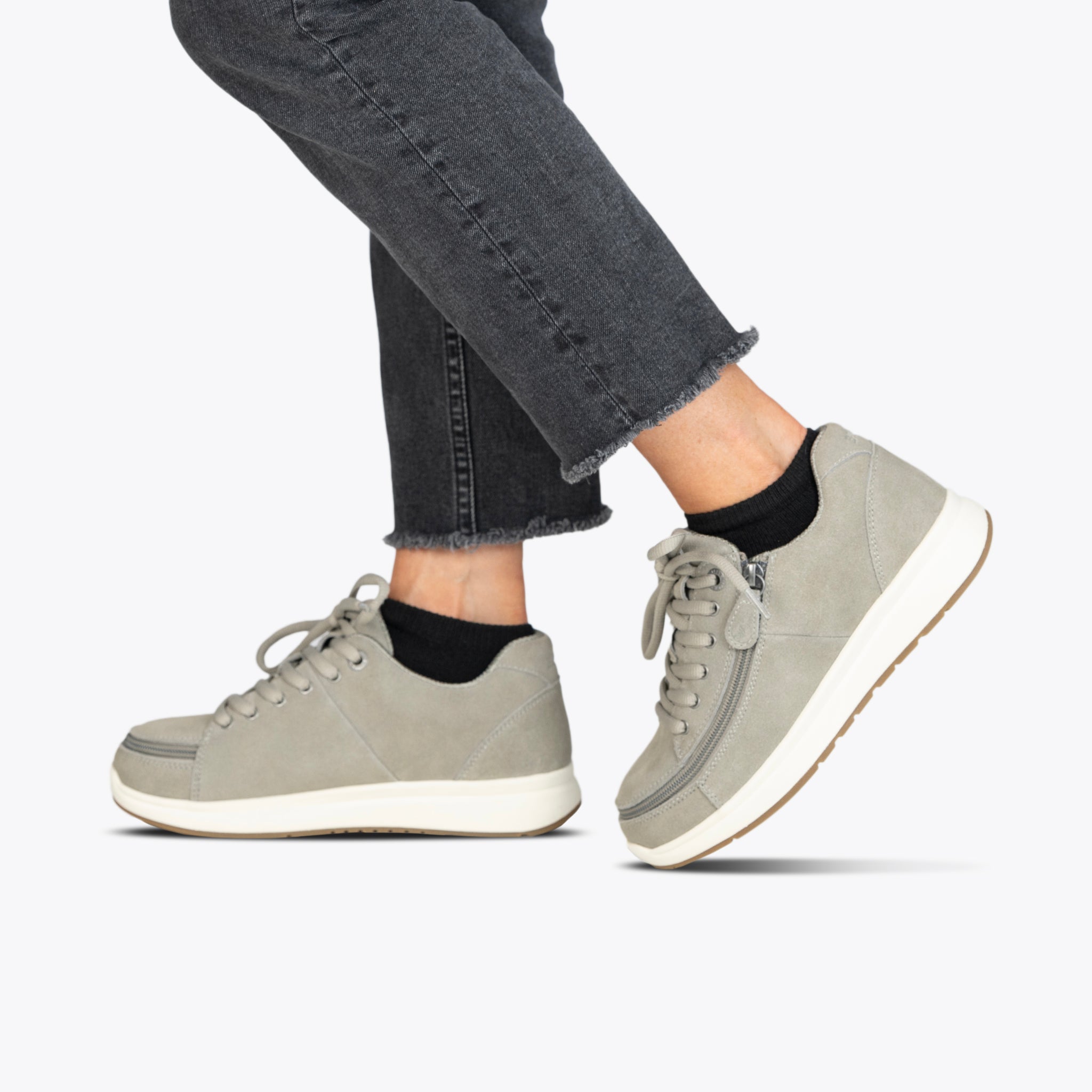 A person wearing Women's Grey Suede BILLY Comfort Plush shoes by BILLY Footwear, with white soles and black socks, pairs them with frayed-hem dark jeans while stepping forward against a plain light background.