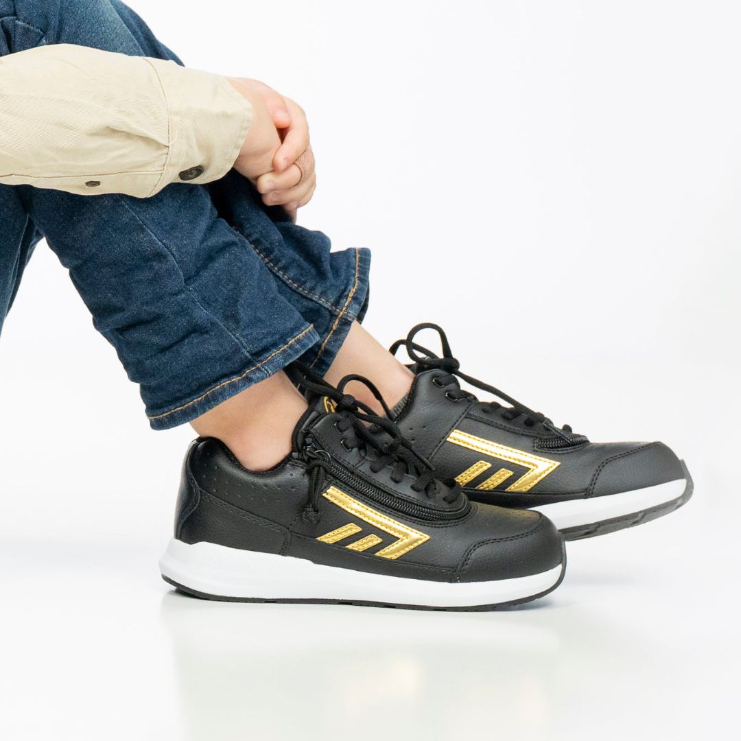 A person wearing blue jeans, a beige shirt, and BILLY Footwear Black/Gold BILLY Goat Hoop shoes sits cross-legged on the floor against a white background.