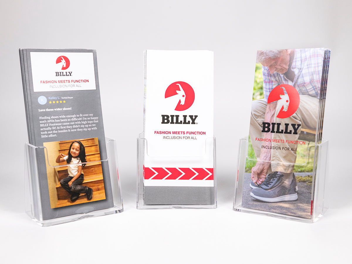 Three clear acrylic brochure holders displaying promotional pamphlets for BILLY footwear. The center brochure shows the red BILLY logo with the tagline “Fashion Meets Function – Inclusion for All.” The left brochure features a review and a photo of a smiling child wearing BILLY shoes while sitting on wooden steps, and the right brochure shows an older man putting on a BILLY shoe.