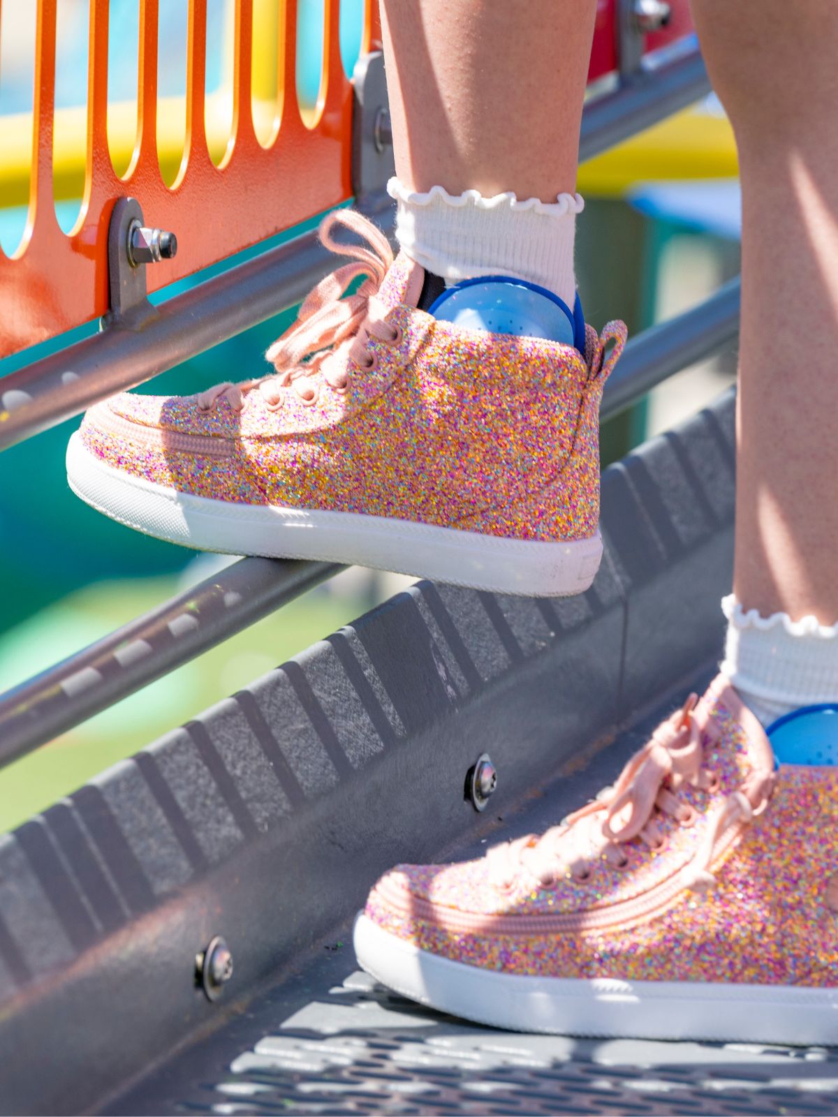 A close-up lifestyle shot of a person's feet wearing pink glitter sneakers with white ruffled socks, standing on a playground metal platform.