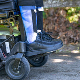 A close-up of lower legs in blue and white orthotic braces, wearing BILLY Footwear Men's Black to the Floor Leather BILLY Comfort Plush shoes and black socks, resting on a motorized wheelchair's footplate outdoors.