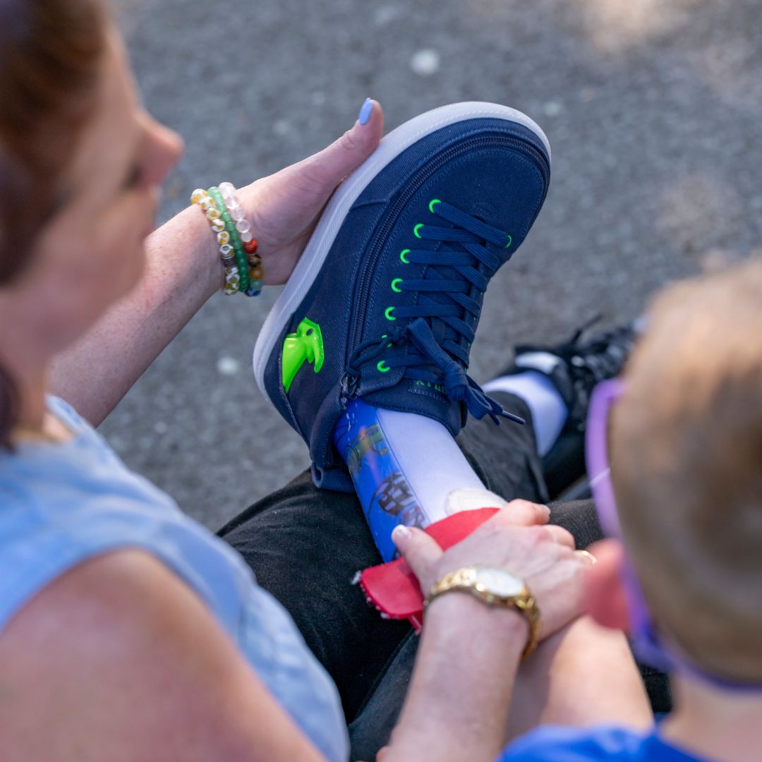 A person helps a child put on the BILLY Footwear Navy/Green Light Ups BILLY Classic D|R High, an AFO-friendly shoe with a wide toe box, over a prosthetic leg as they sit together outdoors. Only their arms and legs are visible.