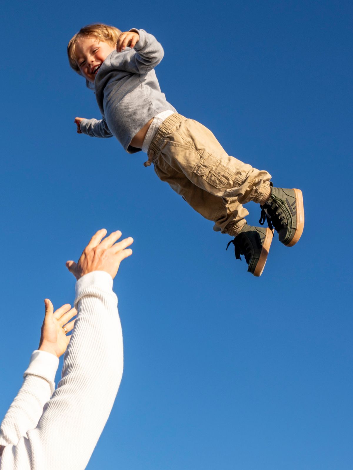 A young child is tossed into the air against a clear blue sky, with an adult's hands reaching up below. The child is wearing a gray long-sleeve shirt, tan cargo pants, and dark sneakers.