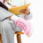 A person in light jeans and bright pink Pink Glitz BILLY Classic Lace High sneakers by BILLY Footwear, featuring a wraparound zipper, sits on a wooden stool writing in a yellow notebook. Only their lower body and hands are visible.
