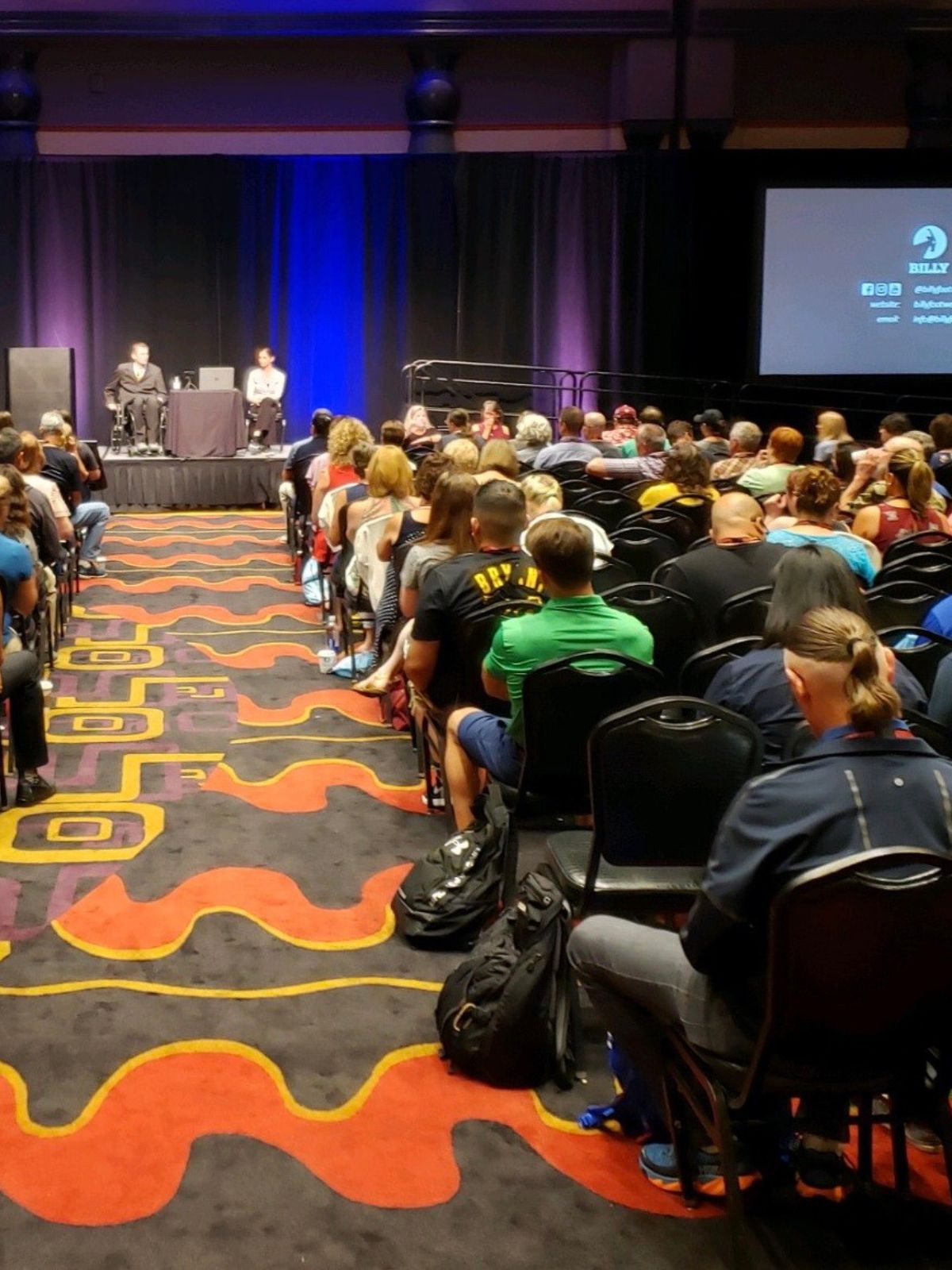 Audience in a conference room with a stage and screen displaying text.