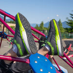 A person wearing SALE - Smoke/Lime Fire BILLY Sport Inclusion shoes from BILLY Footwear with white socks is lying on playground equipment, soles visible. Other people and mountains are in the background.